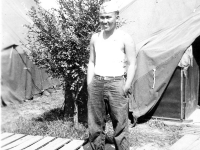 Tokuji Ono standing with hands in pockets on a wooden path in front of tents [Courtesy of Leslie Taniyama] Inscription: Yours Truly Reverse: 8-29-42, 9/7/42