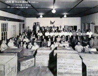 Thomas Higa and wife, Toshiko, are pictured in front center. Photo was taken after packing relief supplies for the victims of the Battle of Okinawa, at the Wailuku Showa Seinen Kaikan (Wailuku Showa Youth Association Hall). Donation crates were sent from Maui to be collected by the Okinawa Clothing Relief Committee at Club 100. (December 15, 1945) [Courtesy of: Thomas Taro Higa Family Collection]